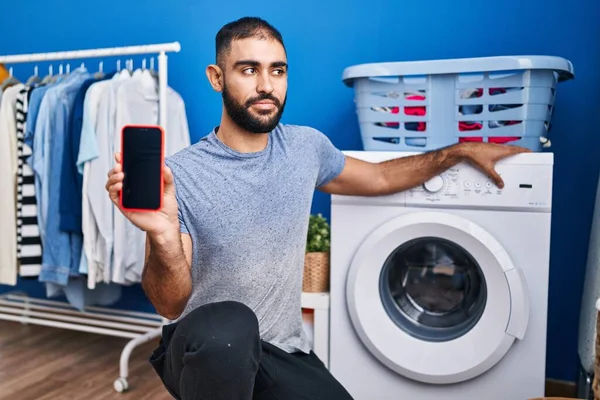 中东男人 留着胡子 露出智能手机屏幕和洗衣机 面带微笑地看着旁边 目不转睛地想着 — 图库照片