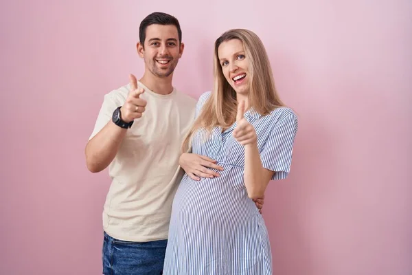 赤ちゃんがピンクの背景に立っていて 楽しそうな顔をしてカメラに指を向けることを期待している若いカップル 良いエネルギーと振動は — ストック写真