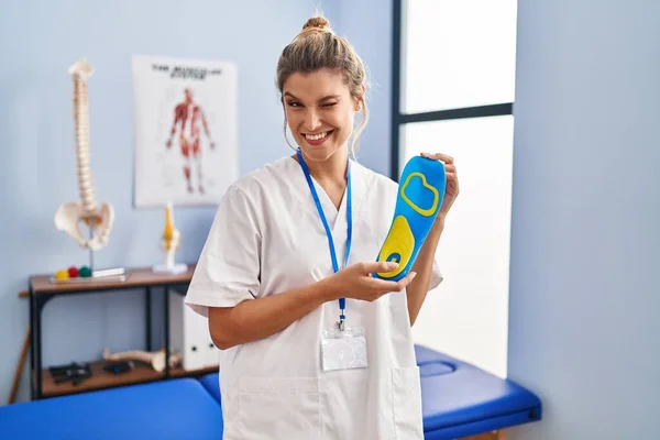 Young woman holding shoe insole at physiotherapy clinic winking looking at the camera with sexy expression, cheerful and happy face.