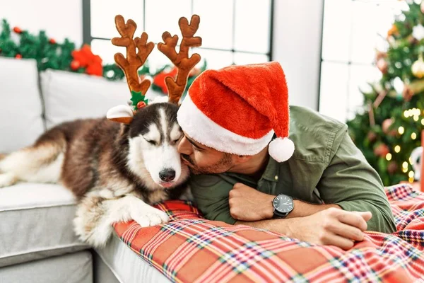 若いですヒスパニック男横たわっソファとともに犬身に着けているトナカイ帽子によってクリスマスツリー ホーム — ストック写真