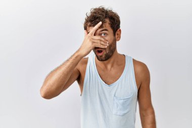 Young handsome man standing over isolated background peeking in shock covering face and eyes with hand, looking through fingers with embarrassed expression. 