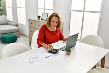 Orta yaşlı, beyaz bir kadın evdeki masada oturuyor..