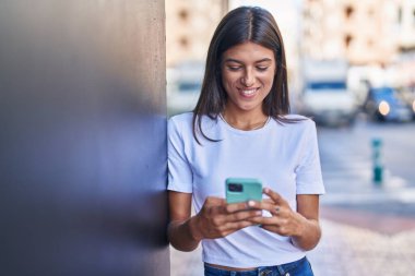 Genç, güzel, İspanyol bir kadın sokakta akıllı telefon kullanarak gülümsüyor.