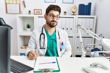 Young man with beard wearing doctor uniform and stethoscope at the clinic smiling looking to the side and staring away thinking. 
