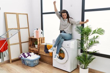 Young hispanic woman listening to music waiting for washing machine at laundry room