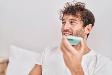 Genç adam akıllı telefonda konuşuyor. Yatak odasında oturuyor.