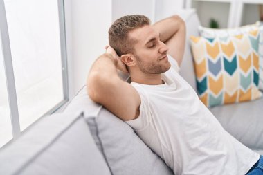 Young caucasian man relaxed with hands on head sitting on sofa at home