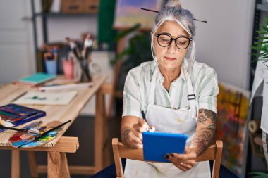 Sanat stüdyosunda dokunmatik ped kullanan orta yaşlı, kır saçlı bir kadın sanatçı.