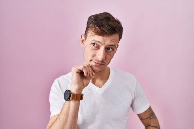 Caucasian man standing over pink background with hand on chin thinking about question, pensive expression. smiling with thoughtful face. doubt concept. 