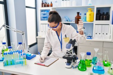 Genç ve güzel İspanyol bilim kadını laboratuarda elinde şişeyle rapor yazıyor.