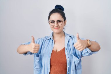 Young modern girl with blue hair standing over white background success sign doing positive gesture with hand, thumbs up smiling and happy. cheerful expression and winner gesture. 