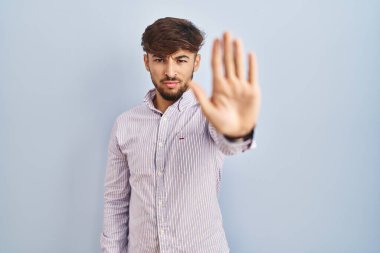 Arab man with beard standing over blue background doing stop sing with palm of the hand. warning expression with negative and serious gesture on the face. 
