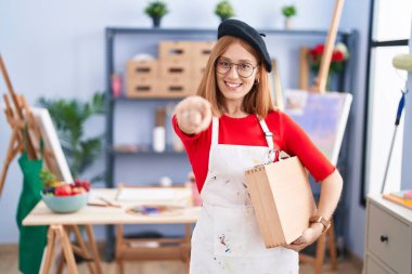 Young redhead woman at art studio holding art case pointing to you and the camera with fingers, smiling positive and cheerful 