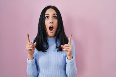 Hispanic woman standing over pink background amazed and surprised looking up and pointing with fingers and raised arms. 