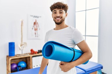 Young arab man patient smiling confident holding yoga mat at clinic