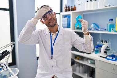 Young hispanic man with beard working at scientist laboratory holding blue ribbon stressed and frustrated with hand on head, surprised and angry face 