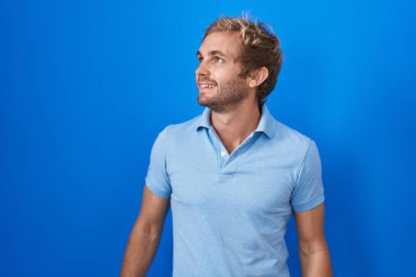 Caucasian man standing over blue background looking away to side with smile on face, natural expression. laughing confident. 