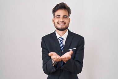 Young hispanic man with tattoos wearing business suit and tie smiling with hands palms together receiving or giving gesture. hold and protection 