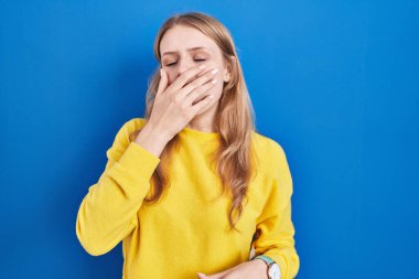 Young caucasian woman standing over blue background bored yawning tired covering mouth with hand. restless and sleepiness. 