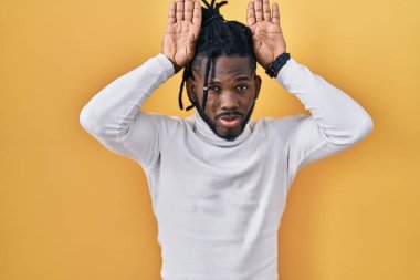 Sarı arka planda balıkçı yaka kazak giyen Afrikalı bir adam avuçlarında alaycı ve şüpheci bakışlarla tavşan kulağı hareketi yapıyor. Paskalya tavşanı konsepti. 