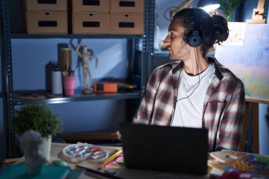 Young hispanic man sitting at art studio with laptop late at night looking to side, relax profile pose with natural face with confident smile. 