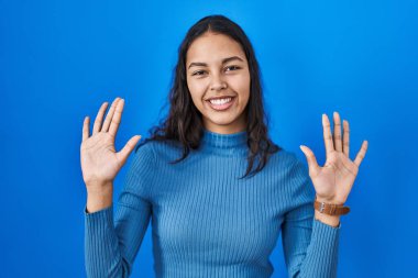 Young brazilian woman standing over blue isolated background showing and pointing up with fingers number ten while smiling confident and happy. 