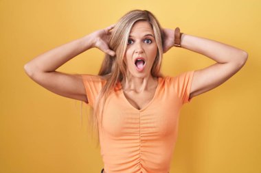 Young woman standing over yellow background crazy and scared with hands on head, afraid and surprised of shock with open mouth 