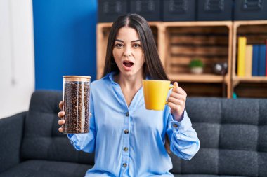 Young brunette woman holding coffee beans and cup of coffee afraid and shocked with surprise and amazed expression, fear and excited face. 