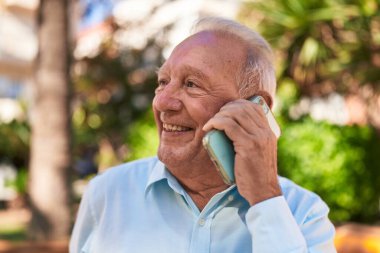 Orta yaşlı, kır saçlı, gülümseyen, kendine güvenen, parkta akıllı telefondan konuşan bir adam.