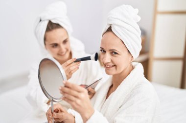 Two women mother and daughter wearing bathrobe applying skin makeup at bedroom
