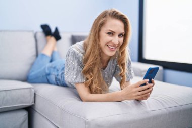 Evdeki kanepede uzanmış akıllı telefon kullanan genç sarışın kadın.