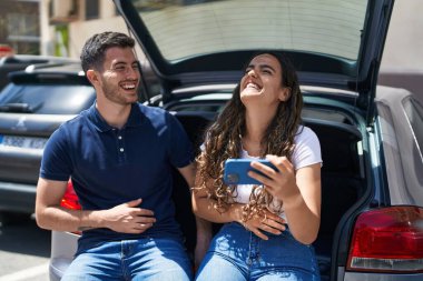 Young hispanic couple sitting on car trunk watching video on smartphone at street