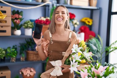 Young woman working at florist shop showing smartphone screen smiling and laughing hard out loud because funny crazy joke. 