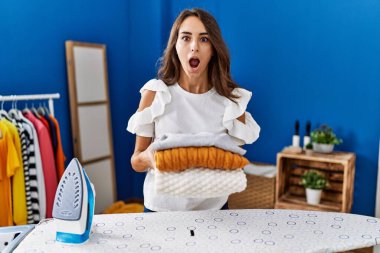 Young hispanic woman ironing clothes at laundry room holding folded sweaters afraid and shocked with surprise and amazed expression, fear and excited face. 