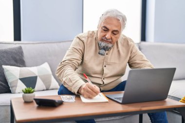 Evde dizüstü bilgisayar kullanan orta yaşlı, kır saçlı bir adam.