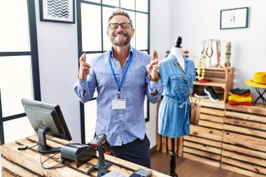 Middle age man working as manager at retail boutique gesturing finger crossed smiling with hope and eyes closed. luck and superstitious concept. 