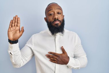African american man standing over blue background swearing with hand on chest and open palm, making a loyalty promise oath 