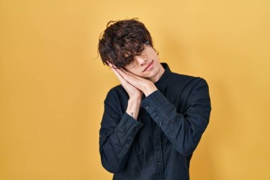 Young man wearing glasses over yellow background sleeping tired dreaming and posing with hands together while smiling with closed eyes. 