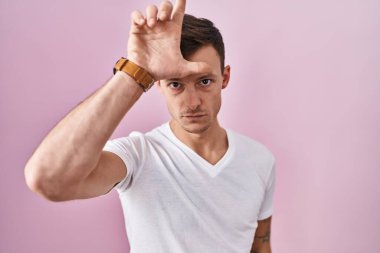 Caucasian man standing over pink background making fun of people with fingers on forehead doing loser gesture mocking and insulting. 