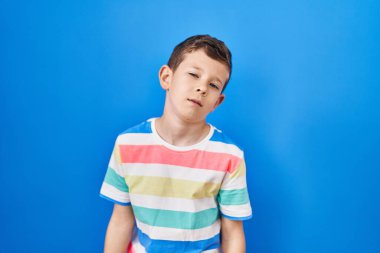 Young caucasian kid standing over blue background looking sleepy and tired, exhausted for fatigue and hangover, lazy eyes in the morning. 