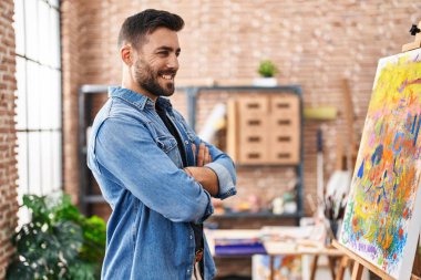 Genç İspanyol adam gülümsüyor. Kollarını kavuşturup sanat stüdyosunda duruyor.