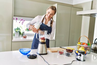 Genç sarışın kadın mutfaktaki miksere süt dökerek gülümsüyor.