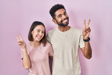 Young hispanic couple together over pink background smiling with happy face winking at the camera doing victory sign with fingers. number two. 