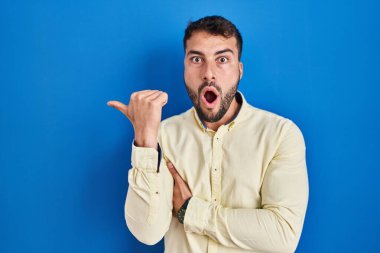 Handsome hispanic man standing over blue background surprised pointing with hand finger to the side, open mouth amazed expression. 