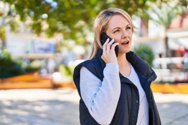 Genç sarışın kadın gülümsüyor. Parktaki akıllı telefondan konuşuyor.