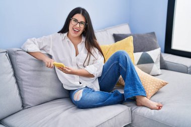 Evdeki kanepede oturan, akıllı telefon kullanan genç İspanyol kadın.