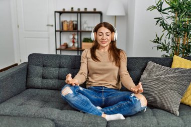 Orta yaşlı beyaz kadın yoga yapıyor ve kulaklık takıyor. Evdeki kanepede oturuyor..