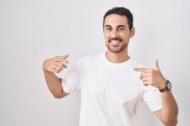 Handsome hispanic man standing over white background looking confident with smile on face, pointing oneself with fingers proud and happy. 