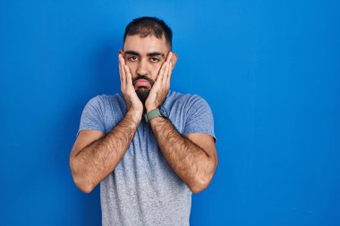Middle east man with beard standing over blue background tired hands covering face, depression and sadness, upset and irritated for problem 
