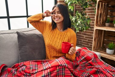 Young hispanic woman sitting on the sofa drinking a coffee at home doing peace symbol with fingers over face, smiling cheerful showing victory 
